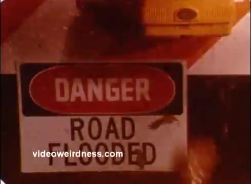 The still features a warning sign reading 'DANGER ROAD FLOODED.' The sign is prominently displayed, likely illustrating a hazardous condition, emphasizing the need for caution. There appears to be water around the sign, reinforcing the message of flood-related dangers. The context is related to a public service announcement by the Red Cross, highlighting emergency preparedness and safety.