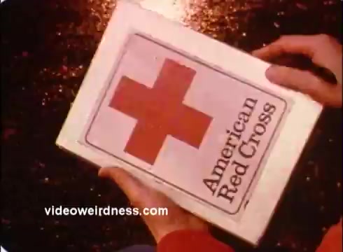 The still features a close-up of hands holding a box displaying the American Red Cross logo, which includes a prominent red cross and the organization's name. The background appears to be slightly blurred, emphasizing the box. The image conveys a sense of urgency and importance, typical of public service announcements aimed at promoting the Red Cross's mission.
