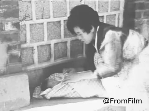 The 16mm still from the 1969 Christian Children's Fund Public Service Announcement depicts a woman bending down towards a child, who is lying on the ground, possibly in a shelter or alleyway. The grayscale image conveys a sense of urgency and compassion, highlighting the theme of care for vulnerable children. The context emphasizes the importance of child sponsorship and support initiatives aimed at improving the lives of disadvantaged youth.