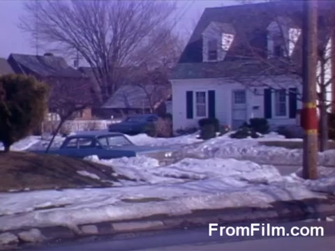 The still from 'Suburbia' (1966 Kodachrome tests) depicts a suburban scene with a modest house featuring a classic gabled roof and white siding. Snow is melting on the ground, suggesting late winter or early spring. A car, possibly from the era, is parked nearby, and the landscape includes bare trees and patches of grass. The overall atmosphere conveys a quiet, residential neighborhood.