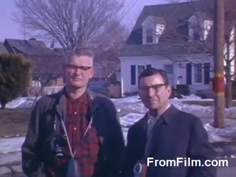The still features two men standing outdoors in a suburban setting, likely in the mid-20th century. They are dressed in formal attire, with one man wearing glasses and a camera slung over his shoulder. In the background, there's a typical suburban house and some trees, indicating a residential neighborhood. The ground shows remnants of snow, suggesting it might be early spring or late winter. The overall color palette reflects the use of Kodachrome film, showcasing vibrant hues typical of that era.