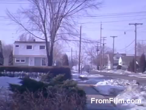 The still from 'Suburbia' features a quiet suburban street in early spring, characterized by remnants of snow on the ground. A leafless tree stands prominently, indicating the transition from winter. The street is lined with power lines and modest homes, showcasing a typical suburban landscape. The colors, captured in Kodachrome, have a distinct vintage quality, contributing to a nostalgic atmosphere.