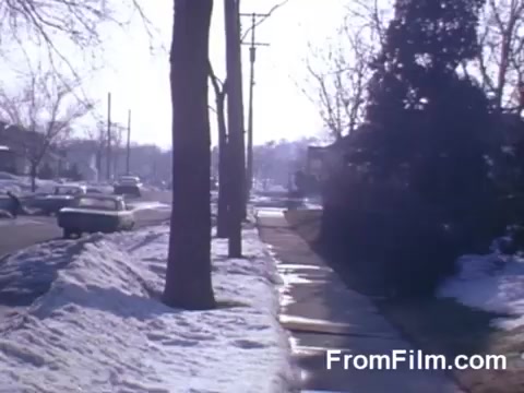 The still from 'Suburbia' features a winter suburban scene, showcasing a snow-covered sidewalk lined with bare trees. Cars are parked along the street, and patches of snow are visible on both the sidewalk and adjacent grass. The atmosphere is bright, illuminated by sunlight, highlighting the serene but stark environment typical of a suburban landscape in winter. The composition reflects a quiet, everyday moment in a suburban neighborhood.