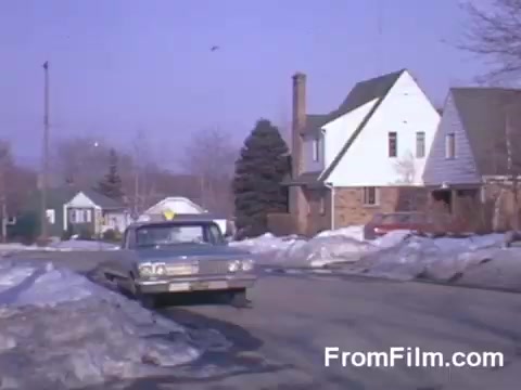 The still from 'Suburbia' (1966) features a serene suburban street scene, showcasing a classic car parked alongside snow-covered areas. The houses are typical of mid-20th century architecture, with visible signs of winter still lingering. The overall color palette, rich in Kodachrome quality, adds a nostalgic feel to the image, hinting at an everyday moment in suburban life during that era.