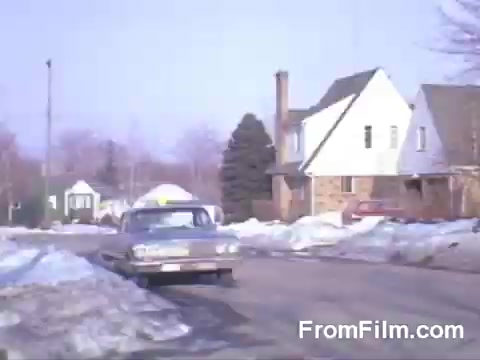 The still features a suburban street scene during winter, with patches of snow lining the roads. A parked car is visible in the foreground, while a residential house with a sloped roof is depicted nearby. The atmosphere conveys a sense of quiet, everyday life in a mid-20th century suburban neighborhood, highlighted by the warm color palette characteristic of Kodachrome film. The image captures a nostalgic, tranquil moment reflecting suburban life.