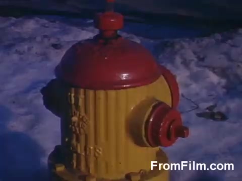 The still features a vibrant yellow and red fire hydrant, prominently positioned amidst a snowy background. The colors of the hydrant pop against the white snow, indicating a cold setting. The image captures a slice of suburban life, highlighting an everyday object that serves a crucial public safety function. The overall tone is nostalgic, reflecting the aesthetic qualities of vintage Kodachrome film.
