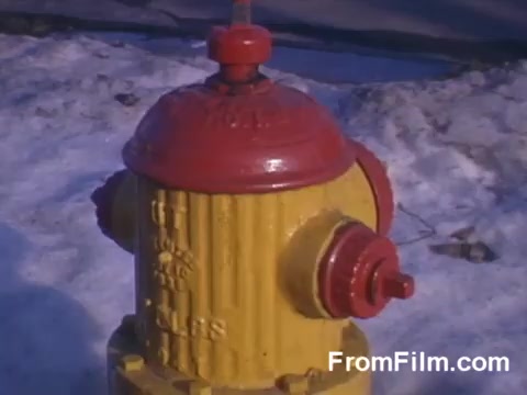 The still features a bright red and yellow fire hydrant, prominently displayed in the foreground against a snowy background. The hydrant's vivid colors contrast sharply with the white, cold surroundings, emphasizing its presence. The image captures a nostalgic and urban feel, typical of mid-20th century American suburbia.