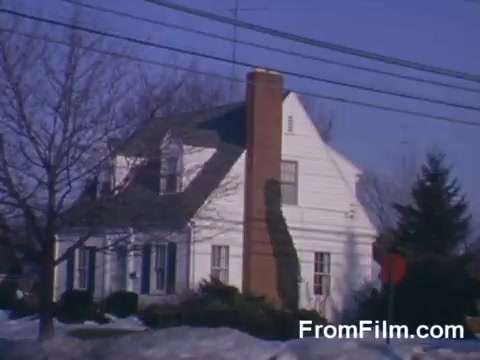 The still features a quaint suburban house characterized by its white exterior, a prominent chimney, and a well-maintained lawn. In the foreground, bare trees are visible, suggesting a winter setting with some snow on the ground. Power lines run across the scene, and a stop sign can be seen nearby, typical of a suburban neighborhood. The color palette is vibrant, a hallmark of Kodachrome film, capturing the essence of mid-20th century suburban life.