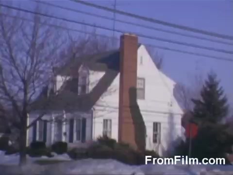The 16mm still features a suburban house typical of mid-20th century American architecture. It shows a light-colored, two-story home with a steep roof, prominent chimney, and a front porch. The surrounding landscape is sparse, with a mix of bare trees and grass, suggesting a winter season. Power lines are visible in the foreground, adding to the suburban setting. The Kodachrome film captures vivid colors typical of that era, highlighting the quaintness of suburban life.