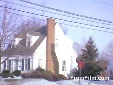 The still features a suburban scene captured in soft Kodachrome hues. A classic white house with a steep roof stands prominently, showcasing traditional architecture. The structure is complemented by a tall brick chimney and flanked by sparse trees, likely indicating winter, as some snow covers the ground. Power lines crisscross the sky, evoking a sense of everyday neighborhood life in the mid-1960s. The overall atmosphere suggests a quiet, residential area typical of suburban landscapes.