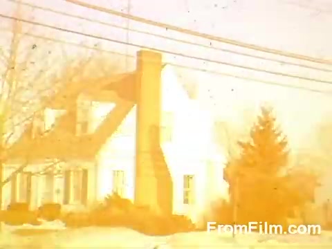 The still from 'Suburbia' features a suburban house prominently displayed against a backdrop of winter scenery. The image has a warm, faded hue characteristic of aging Kodachrome film, giving it a nostalgic quality. Power lines run across the scene, and barren trees are visible, hinting at a quiet residential neighborhood. The overall effect conveys a sense of calm and simplicity typical of mid-20th century suburban life.