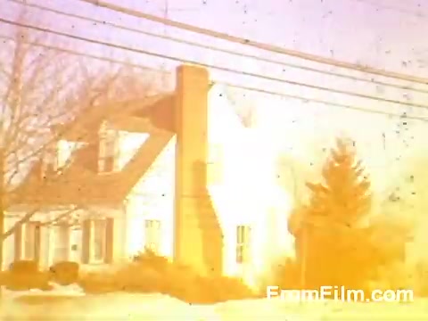 The still from 'Suburbia' features a classic suburban house characterized by its traditional architecture, including a prominent chimney and multiple windows. The scene is bathed in a warm, slightly faded hue typical of 1966 Kodachrome film, suggesting a nostalgic or idealized suburban setting. Power lines crisscross the image, and the surrounding landscape includes some trees and a hint of snow, indicating a cold season. The overall effect evokes a sense of Americana and the tranquil life associated with suburban living during that era.