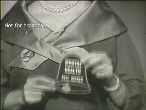 The still from the PSA 'Support Your Mental Health Association' features a woman holding a small bell with 'MENTAL HEALTH' inscribed on it. She is dressed in a stylish outfit typical of an earlier era, complete with a large collar and pearl necklace. The background appears muted, drawing focus to her and the bell, which likely symbolizes raising awareness for mental health. The note 'Not for broadcast' indicates that this is likely an archival image meant for reference rather than public viewing. The overall tone seems serious yet supportive, reflecting the message of promoting mental health awareness.