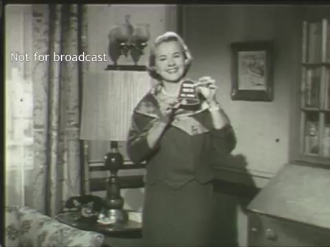 The still from the PSA features a smiling woman in a vintage home setting. She holds up a cup, likely emphasizing a message related to mental health. The background includes a lamp, a floral-patterned couch, and framed artwork, giving it a cozy, domestic feel. The image is black and white, typical of older media, conveying a classic, nostalgic vibe. The text 'Not for broadcast' is visible, indicating it's not meant for public viewing.