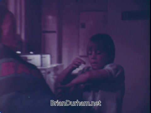 The still from the 'Biz Laundry Detergent' commercial features a young child looking at their arm, possibly questioning a stain or mess. The scene has a nostalgic, slightly faded 16mm quality typical of early 70s commercials, creating a homey atmosphere. A figure in the background, possibly an adult, is partially visible, suggesting a moment of concern or guidance. The overall tone reflects the familial themes often present in advertisements of that era.