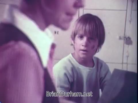 The still shows a young boy with a visible bruise on his face, looking concerned as he interacts with a woman, likely his caregiver or mother. The background features a typical laundry setting with lockers or cubbies. The scene captures a moment of tension, possibly indicating a discussion about cleaning or care, in the context of a commercial for Biz Laundry Detergent from the early 1970s.