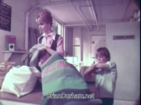The still features a woman and a young boy in a laundromat setting. The woman, dressed in a pink shirt and a vest, is shown handling laundry, while the boy is playfully engaged, likely with a laundry bag. The overall ambiance reflects a casual, domestic scene typical of early 70s advertisements. The focus appears to be on the interactions around laundry, emphasizing the use of Biz laundry detergent.