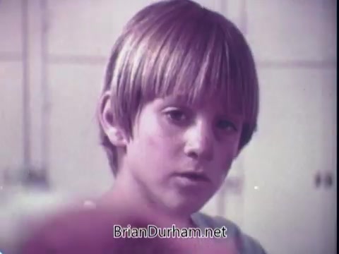 The still from the 'Biz Laundry Detergent' commercial features a young boy with a bowl haircut, gazing intently at the camera. The image has a soft, vintage quality typical of early 70s advertising, with muted colors. The boy's expression suggests curiosity or concern, likely reflecting a moment tied to the product's effectiveness or messaging. The background is minimally detailed, keeping the focus on him.
