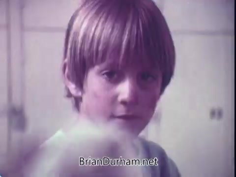 The still features a young boy with a light blond haircut, looking directly at the camera with a neutral expression. The image has a soft, vintage quality typical of 1970s commercials, possibly conveying innocence or curiosity. The background is blurred, focusing attention on the boy, suggesting a domestic setting that aligns with laundry themes.