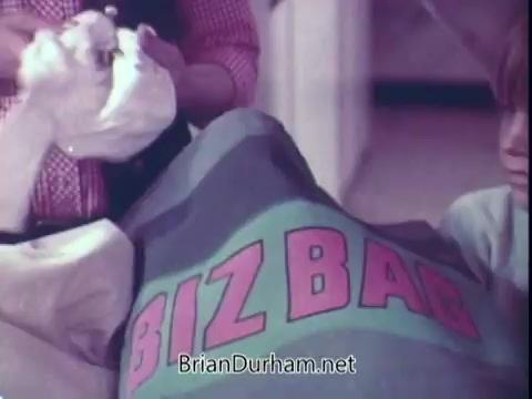 The still features a person holding laundry items while another is seen interacting with a large bag labeled 'BIZ BAG,' indicating that it is a commercial for Biz laundry detergent from the early 1970s. The colors are vibrant, typical of the era's advertising style, showcasing a domestic setting focused on laundry.