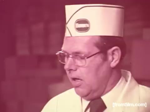 The still features a man in a white uniform and a branded cap, typical of the 1970s food industry. His expression suggests he is engaged in a presentation or demonstration related to Borden American Cheese. The color has significantly faded to a reddish hue, with minimal visible yellows, indicating the age and deterioration of the film. The background appears industrial, possibly a cheese production facility. This captures the essence of vintage advertising from that era.