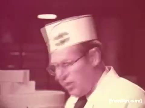 The still from the 'Borden American Cheese' commercial features a man wearing a white coat and a hat with the Borden logo. He appears to be speaking or engaging with the camera, conveying a sense of authority and professionalism in a cheese-related setting. The overall color is heavily faded to red tones, indicative of aging film stock, with minimal remnants of other colors like yellow. The background features light-colored boxes, adding to the commercial's focus on cheese products.