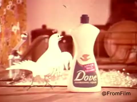 The still from the 1970s Dove Dishwashing Detergent commercial features a whimsical white dove graphic beside the product bottle. The bottle is prominently displayed, emphasizing its dual nature as both a cleaning agent and a gentle hand lotion. The vibrant, retro kitchen background includes floral patterns, enhancing the nostalgic feel of the ad. The caption humorously suggests the product's unique quality, positioning it as a safe and caring choice for dishwashing.