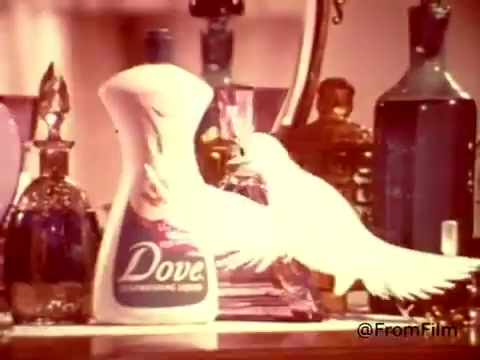 The still from the 1970s Dove dishwashing detergent commercial features a prominently displayed bottle of Dove, designed to evoke a sense of luxury and care. A stylized dove is depicted near the bottle, reinforcing the brand's gentle image. The background consists of elegant, vintage glassware, suggesting sophistication. The tagline humorously contrasts the detergent with hand lotion, emphasizing its mild formula. The overall aesthetic captures the vibrant, colorful style of the era while promoting Dove's unique branding.