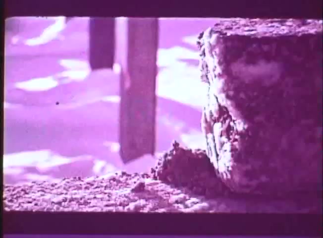 The still from the trailer of *One Day in the Life of Ivan Denisovich* features a close-up of a frozen block of food, likely bread, resting on a snowy surface. The colors have a pinkish hue characteristic of a 16mm print. The scene evokes the harsh conditions faced by the characters in the story, capturing a moment of scarcity and survival in a Soviet labor camp.