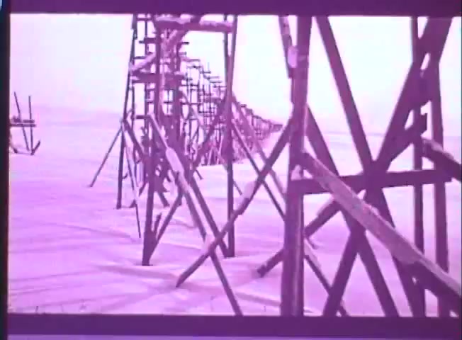 The still shows a stark, desolate landscape characterized by wooden structures resembling scaffolding or old construction supports, set against a snowy backdrop. The color palette has a muted, pinkish tint, typical of 16mm prints, evoking a harsh, cold environment. This imagery aligns with the themes of survival and bleakness present in 'One Day in the Life of Ivan Denisovich.'