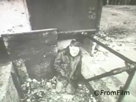 The still from the 'American Institute of Architects PSA - 1969' depicts a somber scene where a man is kneeling in a debris-filled area, suggesting themes of destruction and neglect. The stark black-and-white visual emphasizes the seriousness of the subject matter, likely related to architectural concerns or social issues of the time. This PSA aims to raise awareness about the importance of thoughtful architectural design and its impact on communities.