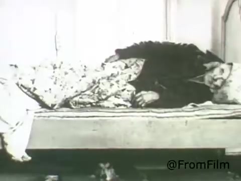 The still from the 'American Institute of Architects PSA -1969' depicts a figure lying on a bed, partially covered by a patterned blanket. The setting appears to be simple, possibly indicative of a domestic environment. The monochrome aesthetic of the 16mm print suggests a focus on the emotional or conceptual aspects of architecture in relation to human experiences, potentially highlighting themes of comfort, neglect, or the human condition.