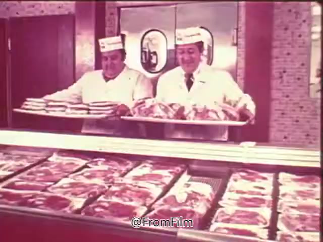 The 16mm still from the 'Swift Pro-Ten Tenderized Beef Commercial' features two butchers in white coats and hats, smiling as they hold trays of packaged beef. The display case in front of them showcases various cuts of meat, emphasizing the product's freshness and quality. The overall atmosphere conveys a friendly and inviting grocery store environment, perfect for a voice-over promoting Pro-Ten beef's availability.