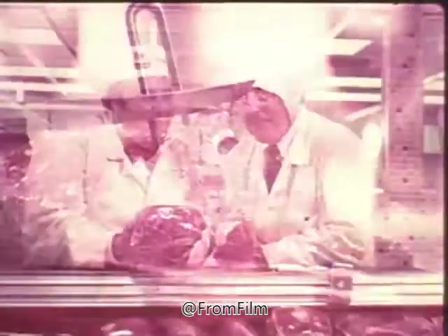 The 16mm still from the 'Swift Pro-Ten Tenderized Beef Commercial' features two men in white lab coats examining a large piece of beef in what appears to be a meat processing facility. The setting has bright overhead lights, creating a sterile atmosphere. This image likely emphasizes the meat's quality and the innovative Pro-Ten tenderizing process. It reflects the commercial's aim to promote the product in a practical, appealing way, with space at the end for a voice-over announcing its availability in local grocery stores.