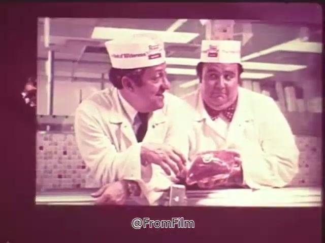The still from the 'Swift Pro-Ten Tenderized Beef Commercial' features two butchers in white lab coats and hats, standing in a meat-processing area. One butcher is gesturing toward a raw cut of beef, while both appear to be engaged in a friendly conversation, likely promoting the tenderness of Pro-Ten beef. The backdrop includes tiled walls typical of a butcher shop. The commercial is positioned to conclude with a voice-over announcing availability at local A&P grocery stores.
