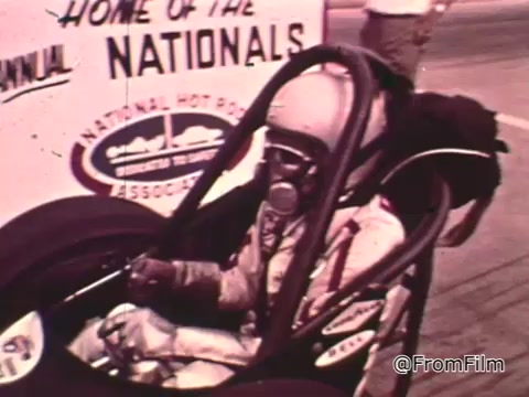 The still depicts a classic drag racing scene featuring a race car and a driver in an enclosed helmet, seated in a dragster. The background includes signage indicating the event is related to the National Hot Rod Association (NHRA) Nationals, likely in the 1960s.   The car's design, typical of that era, is characterized by a sleek, long body, common in drag racing vehicles of the time. Identifying the specific Dodge model requires more detail, but Dodge was known for producing powerful vehicles during that period, often used in drag racing. Without additional specifics, pinpointing an exact year for the Dodge shown is challenging.