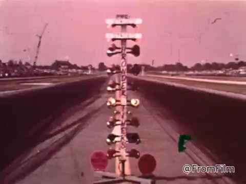 The still features a drag strip with a light tree used for timing races, set against a warm, tinted sky typical of the 1960s. This visual context suggests a focus on the intensity and excitement of drag racing.   As for the Dodge mentioned, without more specific details about the model or design elements visible in the image, pinpointing the exact year is challenging. Dodge vehicles from the 1960s, particularly those associated with drag racing, often include models like the Dodge Charger or Dodge Dart, but identification would require more context.
