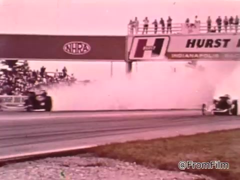 The still from 'Dragsters and Cars' features a drag racing scene, showcasing vehicles racing down a track with smoke billowing from the cars. The setting includes a crowd watching from a grandstand and a signage displaying the NHRA (National Hot Rod Association) logo, indicating a professional drag racing event.  As for the Dodge in question, identifying the exact year based solely on the image is challenging without additional context or details about the model. Generally, Dodge cars from the 1960s that are popular in drag racing include models like the Dart and Charger.