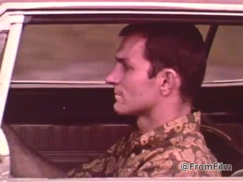 The still depicts a driver inside a car, likely a Dodge model from the 1960s, given the context of the PSA featuring Don Garlits, a notable drag racer of that era. The driver wears a patterned shirt, focusing on the road ahead. The visual emphasizes car maintenance and performance, integral to succeeding in drag racing. For a specific year of the Dodge, further details or identification would be needed.