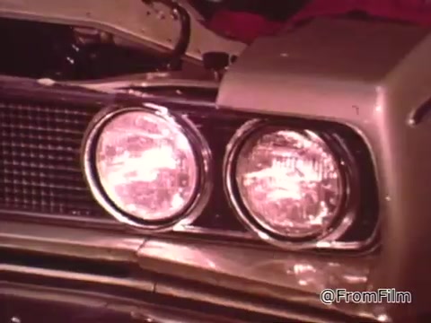 In the 16mm still from 'Dragsters and Cars,' the focus is on the front of a Dodge vehicle, specifically highlighting its dual headlights and grille. The scene reflects the era of 1960s drag racing, emphasizing car maintenance and performance. As for the year of the Dodge, it likely pertains to models from the mid to late 1960s, which were popular during that period of drag racing.