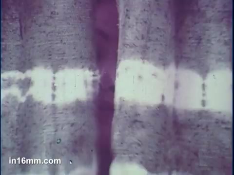 The still shows a piece of fabric with a tie-dye pattern, featuring distinct stripes of color. The image has a soft focus, creating a dreamy effect. A face is partially visible behind the fabric, suggesting a playful or mysterious element typical of commercial aesthetics. The overall color scheme leans towards warm tones, enhancing the artistic vibe of the tie-dye design.