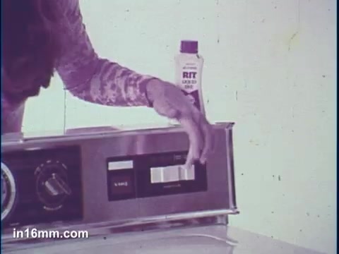 The still features a close-up of a hand adjusting a control on a washing machine, with a bottle of Rit dye prominently displayed beside it. The setting is likely a laundry room, and the overall aesthetic reflects a vintage style commonly seen in 16mm commercials. The focus suggests the application of the dye in a tie-dye process, emphasizing the product's use in crafting colorful fabrics.