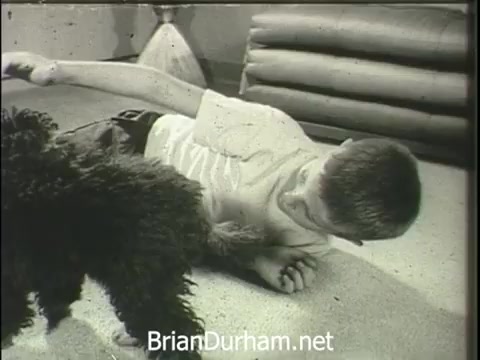 The still from 'Wheeling Electric Company - Electric Water Heaters' features a playful interaction between a boy and a dog on a carpeted floor. The boy, lying on his side, appears engaged and amused, while the dog is curiously approaching him. The setting captures a warm, friendly atmosphere, likely intended to convey the comfort and reliability of electric water heaters in a home environment. The humorous comparison between the dog and the utility of the water heater emphasizes its convenience and friendly nature.