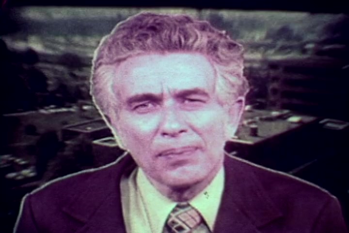 The still from 'Drug Abuse: Some Comments from the Morgue' features Tillman Hahn from the Philadelphia Medical Examiner's office. He appears in a serious manner, addressing the camera. The background depicts a faded urban landscape, suggesting a somber tone. The color palette is muted, typical of 1970s film, emphasizing the film's anti-drug message through Hahn's expression and demeanor. The overall mood is one of concern and gravitas related to the topic of drug abuse.