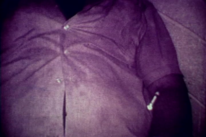 The still depicts a close-up of an individual's arm, resting on a surface. The person is wearing a button-up shirt, partially buttoned, revealing evidence of drug use—specifically, a syringe lying near the forearm. The image has a muted, slightly discolored quality typical of 16mm film, aligning with the film's serious anti-drug message from the 1970s. The context suggests a stark reminder of the consequences of drug abuse.