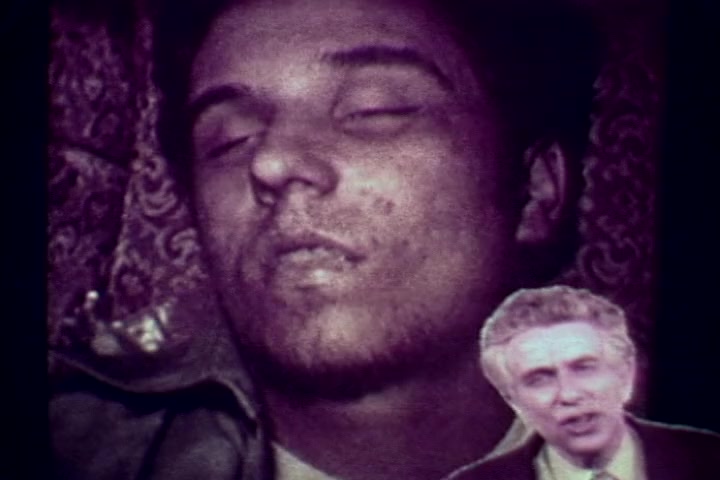 The still captures a somber scene from the anti-drug film 'Drug Abuse: Some Comments from the Morgue.' It features a deceased young man lying on a patterned surface, with his eyes closed and a grim expression. Overlaid is a smaller image of Tillman Hahn, a narrator from the Medical Examiner's office, providing commentary. The film's stark visuals aim to highlight the tragic consequences of drug abuse, reflecting the serious tone of the 1970s public health messaging.