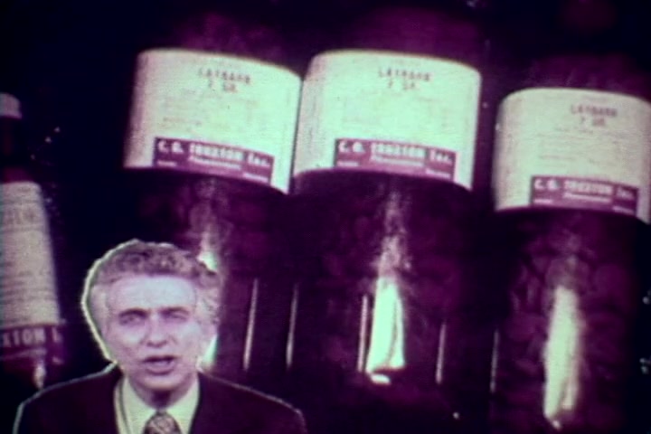 The still features Tillman Hahn speaking directly to the camera, set against a backdrop of large bottles labeled 'LATARG' from C.B. Therapeutics. The contrast of his serious expression with the ominous presence of prescription drugs underscores the film's anti-drug message from the 1970s, likely addressing the dangers of substance abuse. The image has a vintage quality typical of 16mm prints, enhancing its historical context.