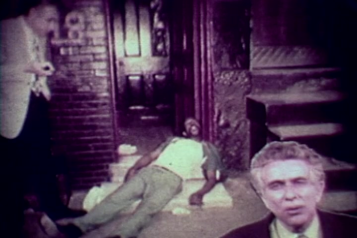 The still from 'Drug Abuse: Some comments from the morgue' depicts a grim scene on urban steps. A deceased individual lies on the ground, visibly affected by drug use. In the background, a man in a light-colored suit appears to observe the situation, while Tillman Hahn, a figure from the Medical Examiner's office, addresses the camera. The image captures a stark and somber moment, reflecting the film's anti-drug message in the context of the 1970s.