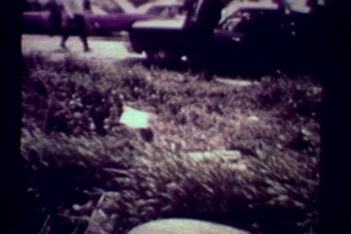 The still from 'Drug Abuse: Some Comments from the Morgue' depicts a scene that appears to be a street or parking lot, with cars in the background and some individuals walking by. The foreground features overgrown grass and litter, suggesting neglect and a possibly urban environment. The color is muted, characteristic of 16mm film, lending a somber tone. This visual context aligns with the film's anti-drug message, emphasizing the grim realities of drug abuse.