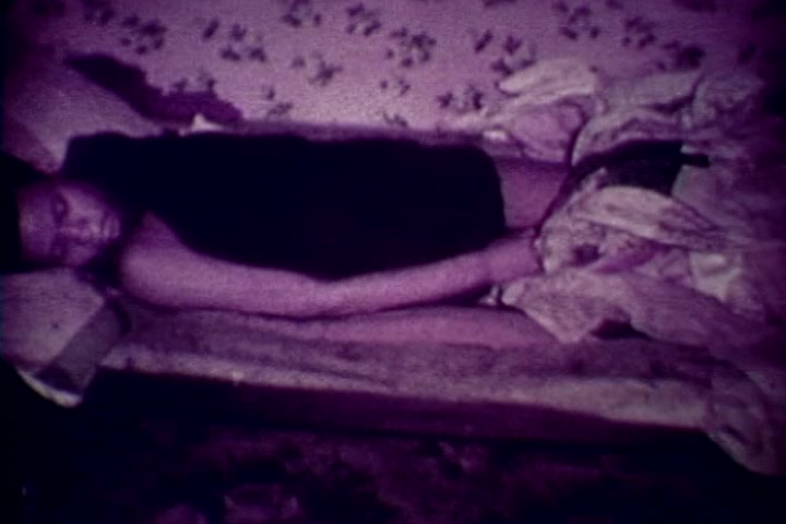 The still features a lifeless figure lying on a makeshift bed, surrounded by crumpled bedding. The scene is dimly lit with a purplish hue, contributing to a somber and unsettling atmosphere. The setting appears to be informal, suggesting a stark reality of drug abuse consequences, likely serving as a powerful visual commentary in the anti-drug film. The focus is on the impact of addiction and mortality, reflective of the film's message.